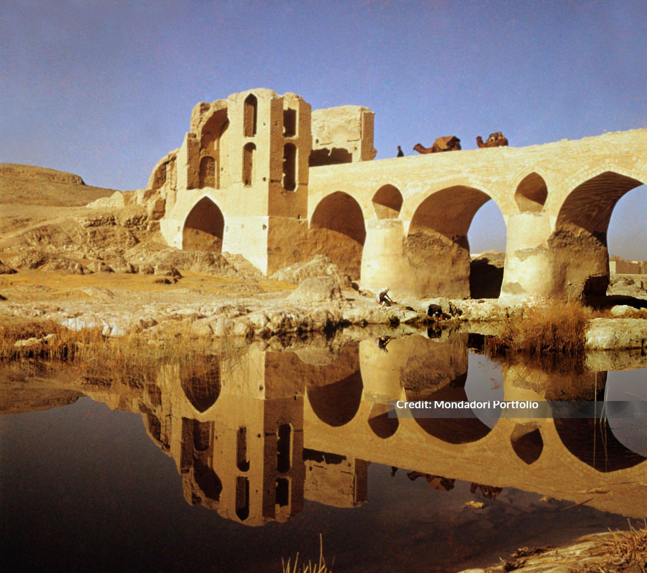 A caravan of camels crossing the Sharestan bridge reflected in the water, Iran, 1960s.