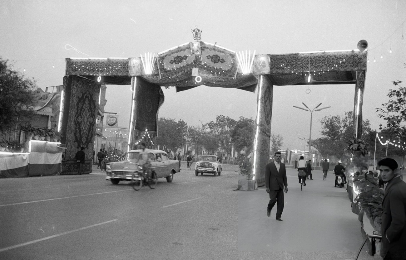 Cars driving under arches decorated with Persian carpets, neon lights, and flags on a grand avenue for the 42nd birthday of Shah Mohammad Reza Pahlavi, Tehran, 1960.