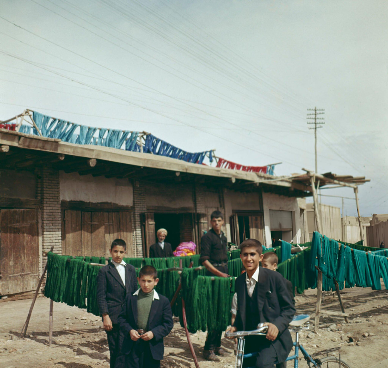 Local boys congregate in front of carpet wools being hung out to dry after dyeing in various colors at a workshop on a street in Iran, 1963.