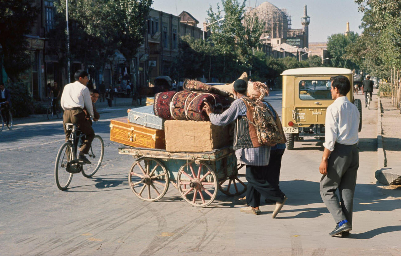 A porter pushing a cart loaded with luggage and carpets across a road in Mashhad, the capital of Razavi Khorasan Province in Iran, 1965.