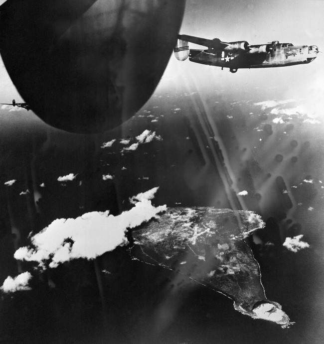 While U.S. B-24 (Liberators) soar high above the Japanese airbase on Iwo Jima, bombs dropped from the planes explode on the island, 1944.