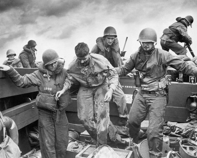 Wounded when Jap fire made a direct hit on an Amtrac, a Marine is transferred by Coast Guardsmen to a landing craft off the flaming shore of Iwo Jima, Japan on D-Day, 1945.