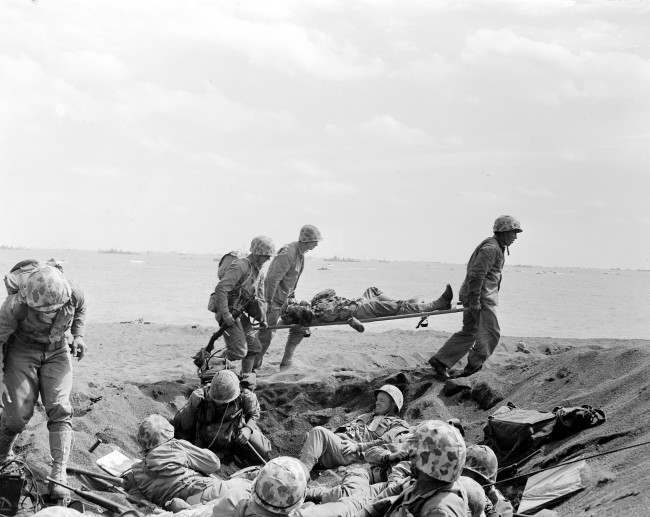 Corpsmen carry a wounded Marine on a stretcher to an evacuation boat on the beach at Iwo Jima while other Marines huddle in a foxhole during the invasion of the Japanese Volcano Island stronghold, 1945. A search team is on the island looking for a cave where the Marine combat photographer who filmed the famous World War II flag raising is believed to have been killed in battle nine days later, military officials said Friday, June 22, 2007.