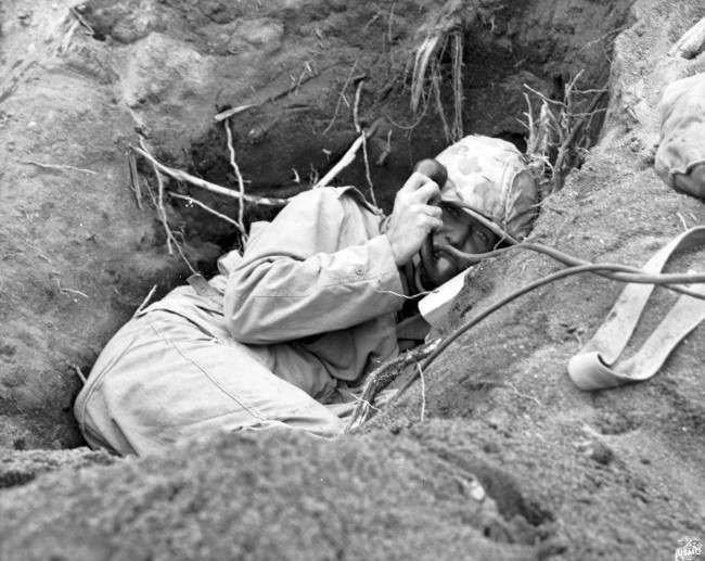 This is a photo of a U.S. Marine communicator, burrowed in a shallow foxhole, calling for artillery support to silence enemy mortars in Iwo Jima, Japan, during World War II, 1945.