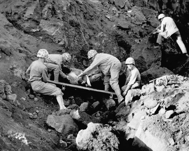 Marine stretcher bearers place a Japanese prisoner on a litter to be carried to the rear, while another Marine covers a cave entrance with his automatic rifle at northern Iwo Jima, Japan, 1945.