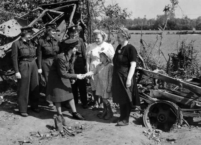 Members of the first WAAF contingent to arrived are greeted by French people in Normandy, 1944. In the back ground is a burnt-out German motor vehicle.