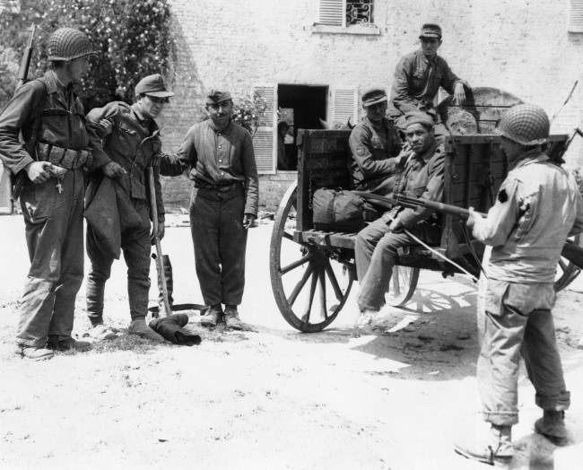 An American soldier and a French peasant assist a wounded Nazi prisoner into a cart in which he and others captured in a small French village, in Normandy, 1944, were transported back to the beach en route to England and internment.