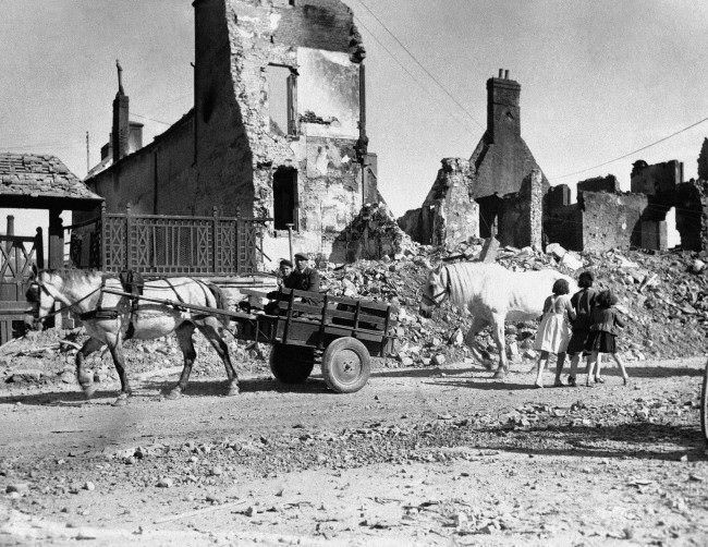 Natives of a French village shattered by the Normandy invasion fighting move down a road past ruined buildings, 1944.