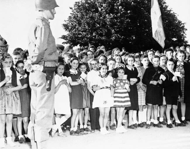 French children of the Normandy town of Grandcamp, France, some carrying American flags and others dressed in patriotic costumes, join American occupation troops in celebrating the American Independence Day, 1944.