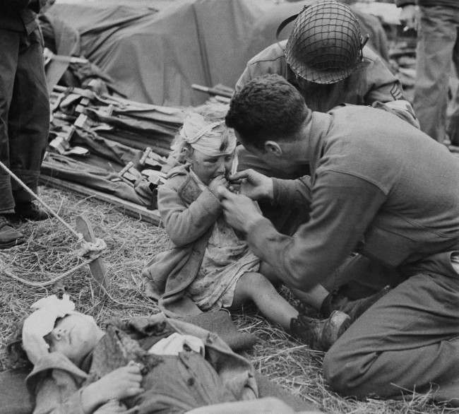 American Army medical corpsmen try to assuage the grief of a little French girl with a gift of candy in France, 1944. Her head is bandaged and face swollen. Another child lies in front with his head bandaged. Both youngsters are evidently under the effects of the terror of Normandy.