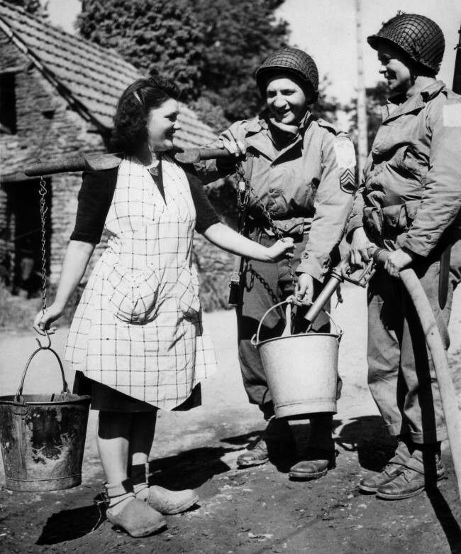 Two American soldiers, Staff Sgt. Bernard Dargols, New York, and Tech., 5/c William L. Stanley, Houston, Texas, help a French farm girl fill her water pails from an army water purification unit during a quiet moment somewhere in the Normandy beachhead area of France, 1944.