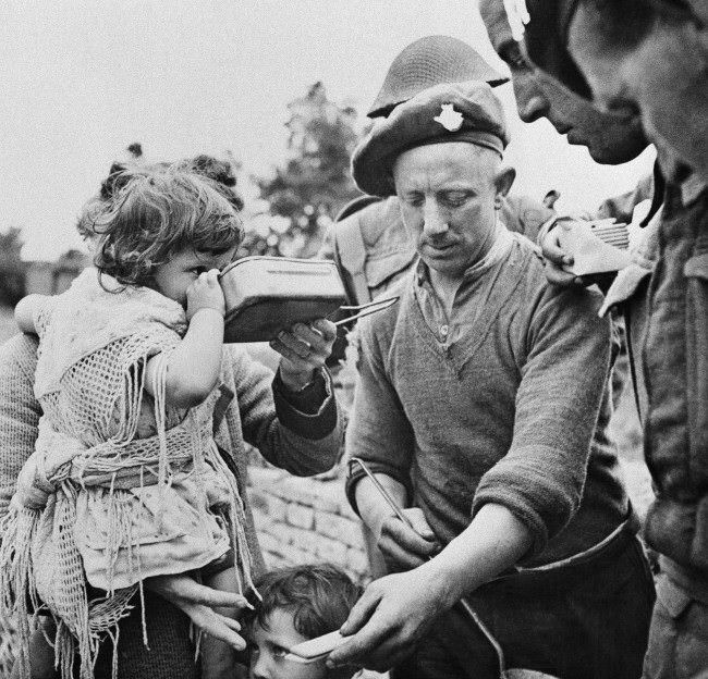 French refugees, including women and children, gather around British soldiers at a Civil Affairs Feeding Center in the Normandy beachhead sector who are supplying hot food, 1944.