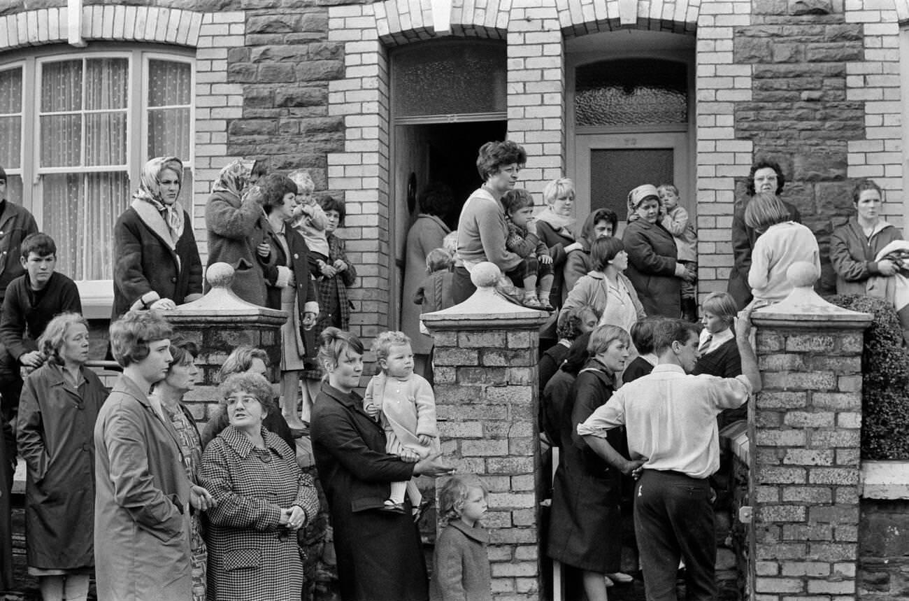 Local villagers, families, young and old look on in horror towards the school and scene of devastation, waiting for news, 1966.