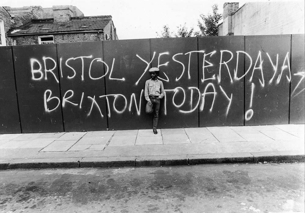 A young man standing in front of a fence with a graffiti message of racial unrest, 1980.