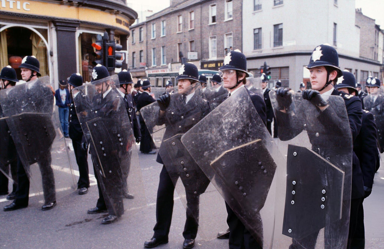 British police officers in uniform and sheltered behind shields in the street during the Brixton racial riots in London, 1981.