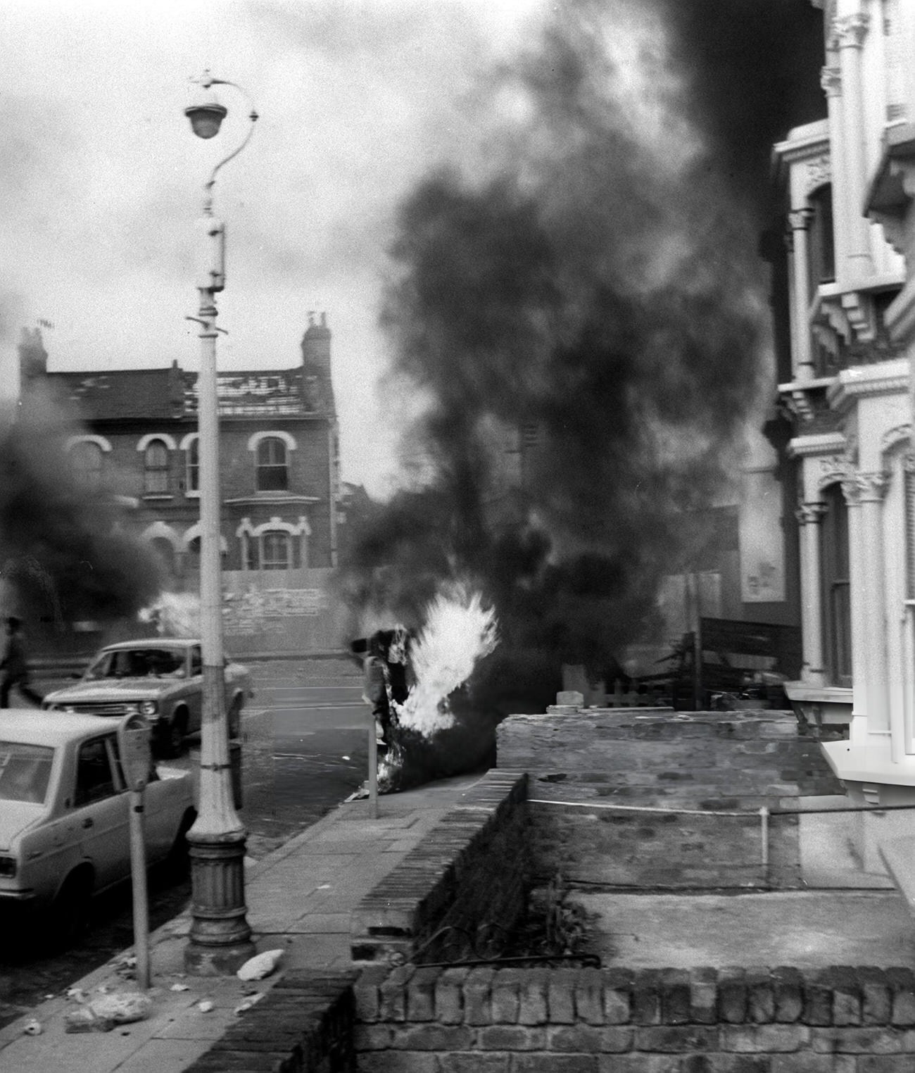 A blazing vehicle on a street at Brixton, South London, 1981.