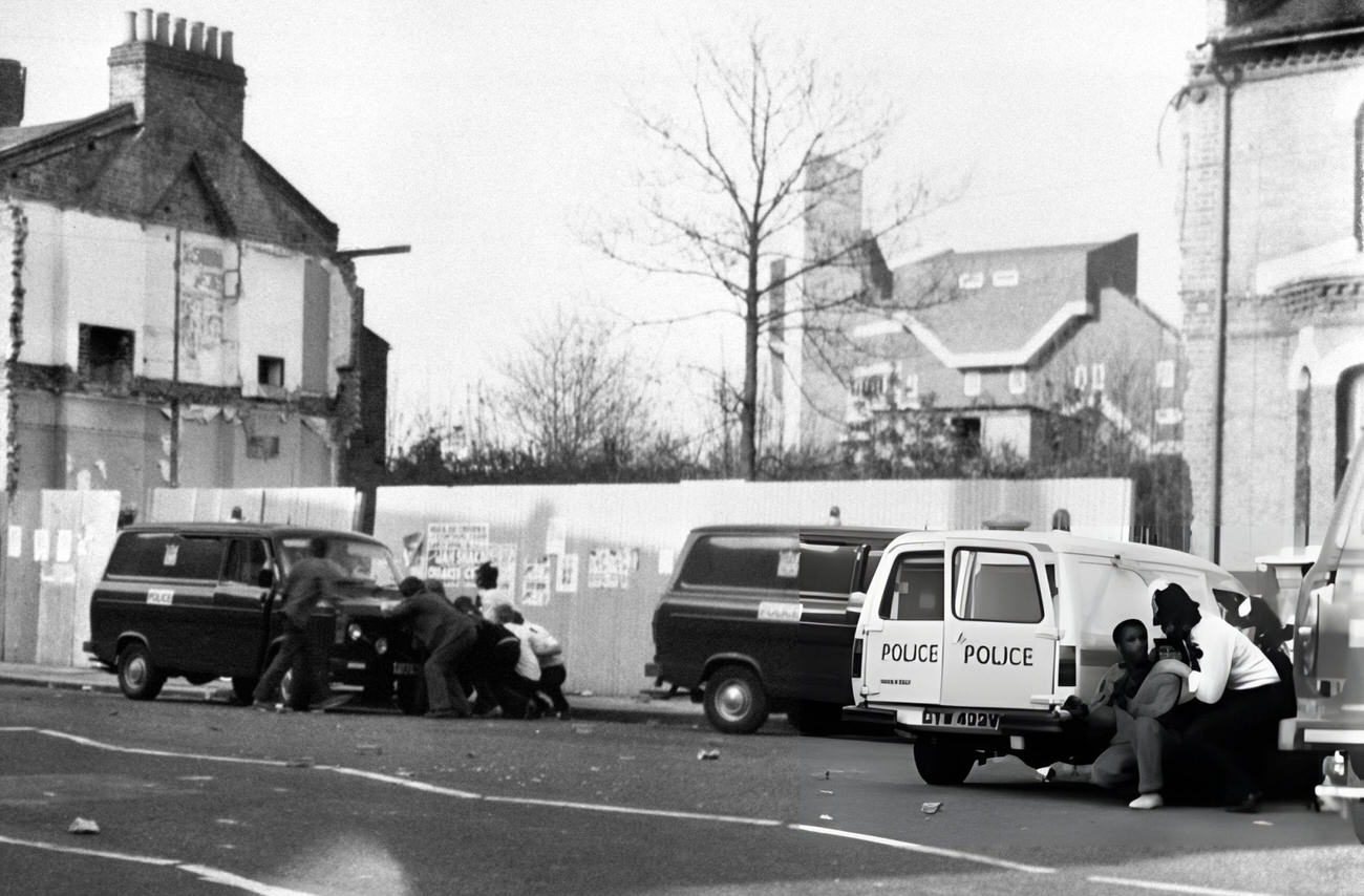 Police Officers shelter behind one of their vans as youths attack another vehicle. Ten Police Officers were injured in Brixton, South London during renewed fighting with black youths. At least one police vehicle was set on fire, 1981.