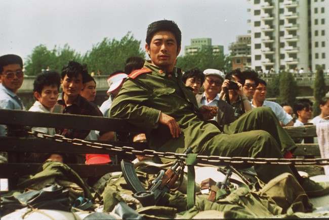 A People’s Liberation Army Soldier sits alone with his weapons Sunday after his convoy was stopped by demonstrators seen in the background.