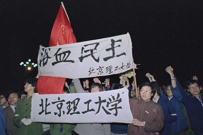 Students from Beijing University of Engineering carry a banner, top, reading ‘Democracy, Bathed in Blood,’ as they march into Beijing’s Tiananmen Square to join tens of thousands of other students early on Saturday, April 22, 1989. The students plan to spend the night in the square and wait for the funeral of Hu Yaobang later in the day.