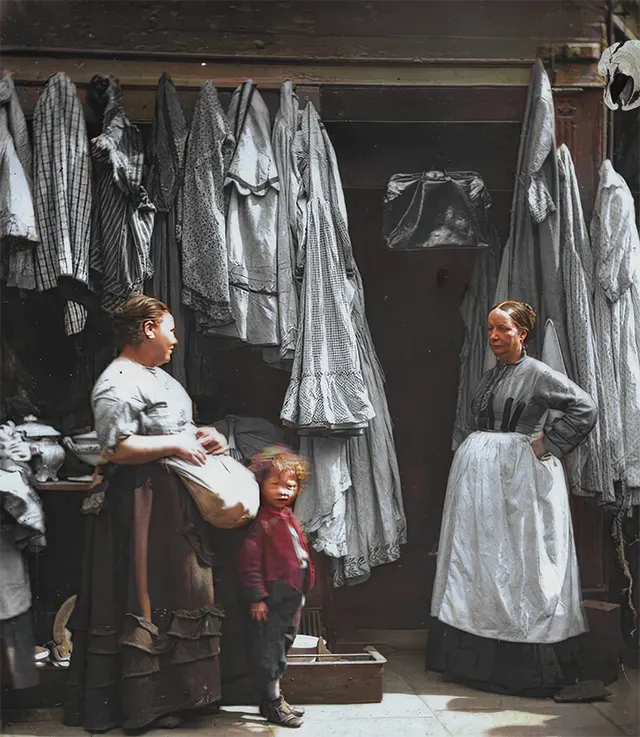 Two women and a child at a secondhand clothing shop in St. Giles, 1890s.