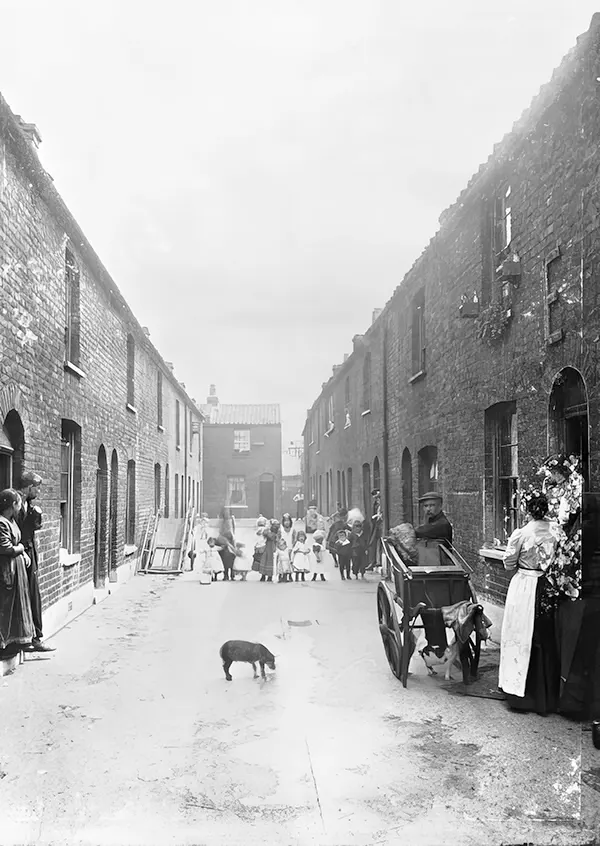 A man selling meat for cats walks down the street of a London slum. The man pushes a cart with a cat strolling between the wheels. 1900s.