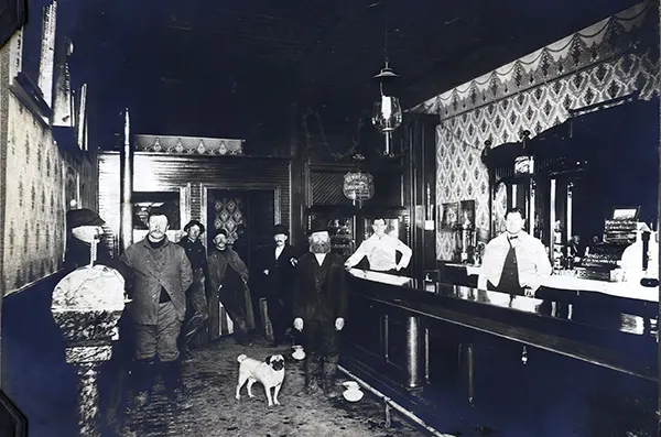 Several men, and a dog named Peggie, stand in John Powell’s Saloon in Plainfield, Illinois, 1910s.