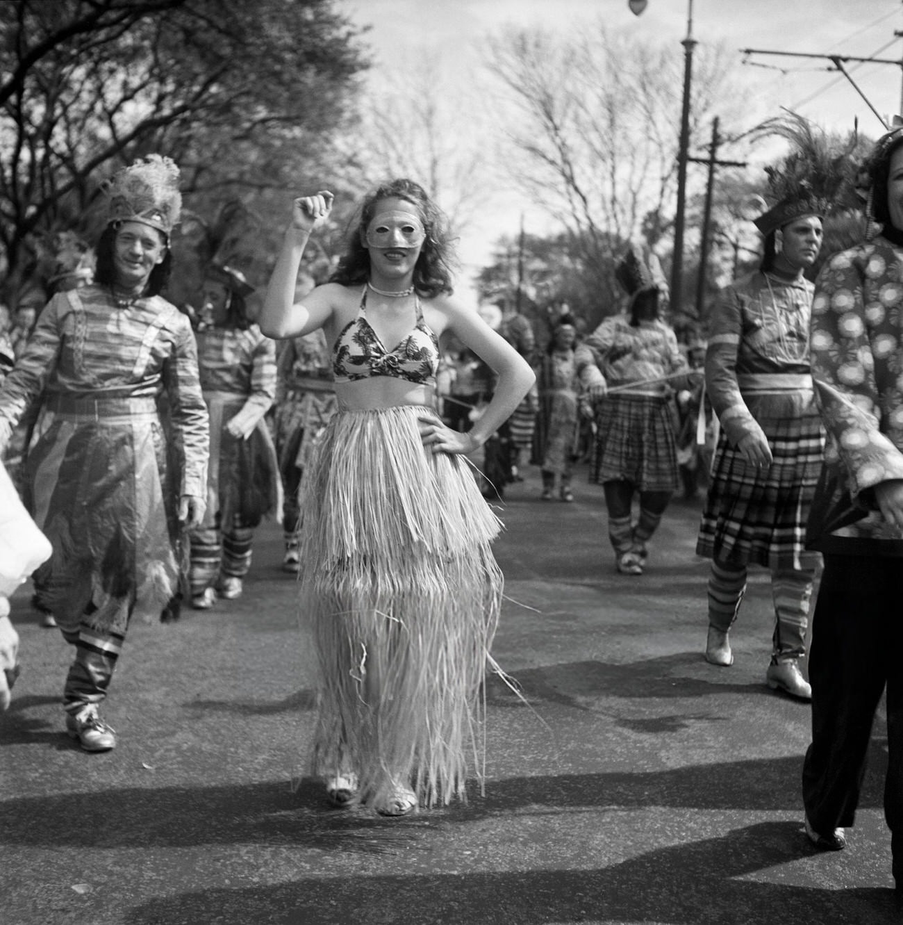 A Woman Dances in a Hula Skirt in a Mardi Gras Parade Amidst Men in Costumes, 1950s.