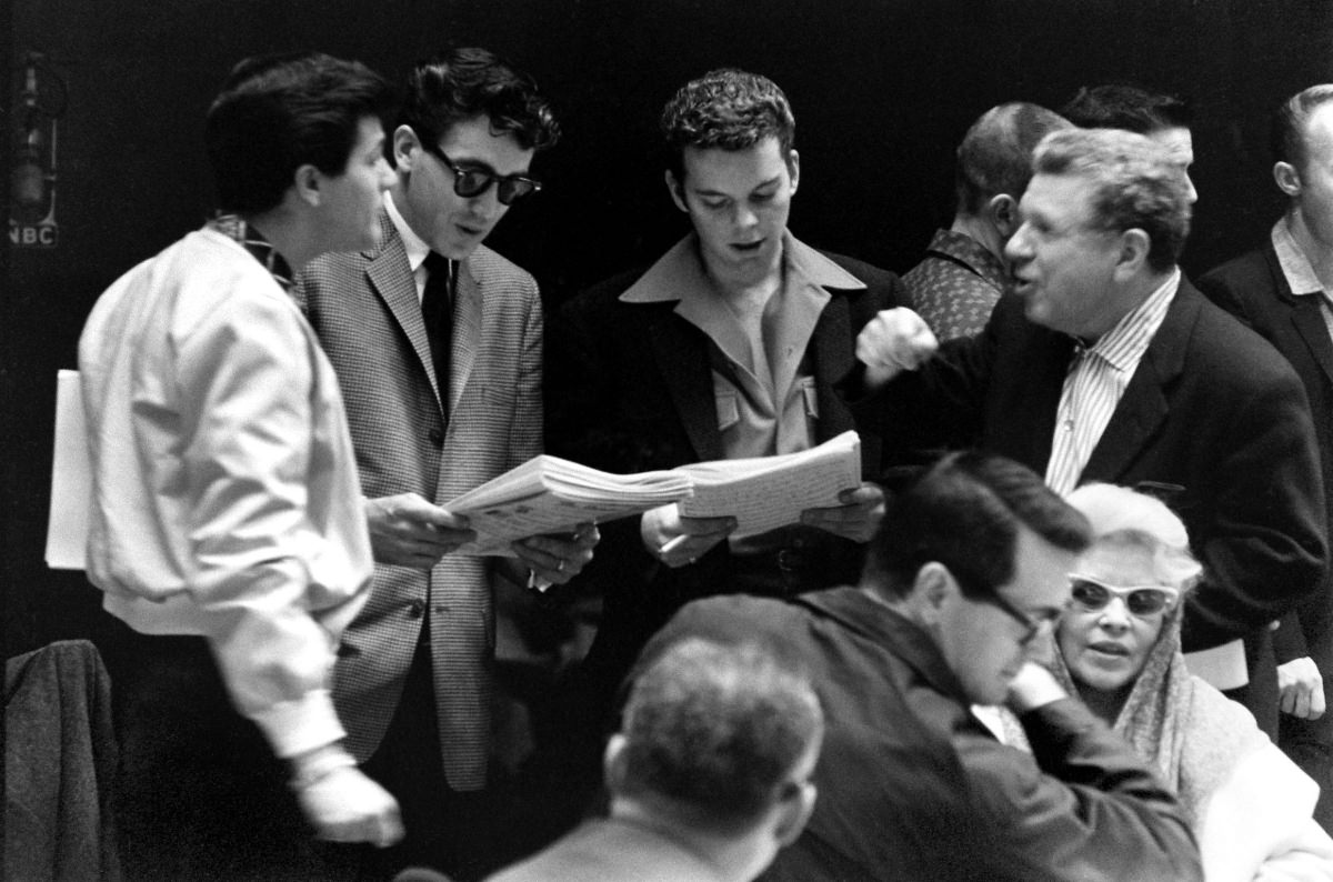 Russ Tamblyn (center, in dark jacket and shirt with huge lapels), 23-year-old Best Supporting Actor nominee for Peyton Place, stands in a group with other unidentified young actors; to the lower right of the frame are Rock Hudson and Mae West.