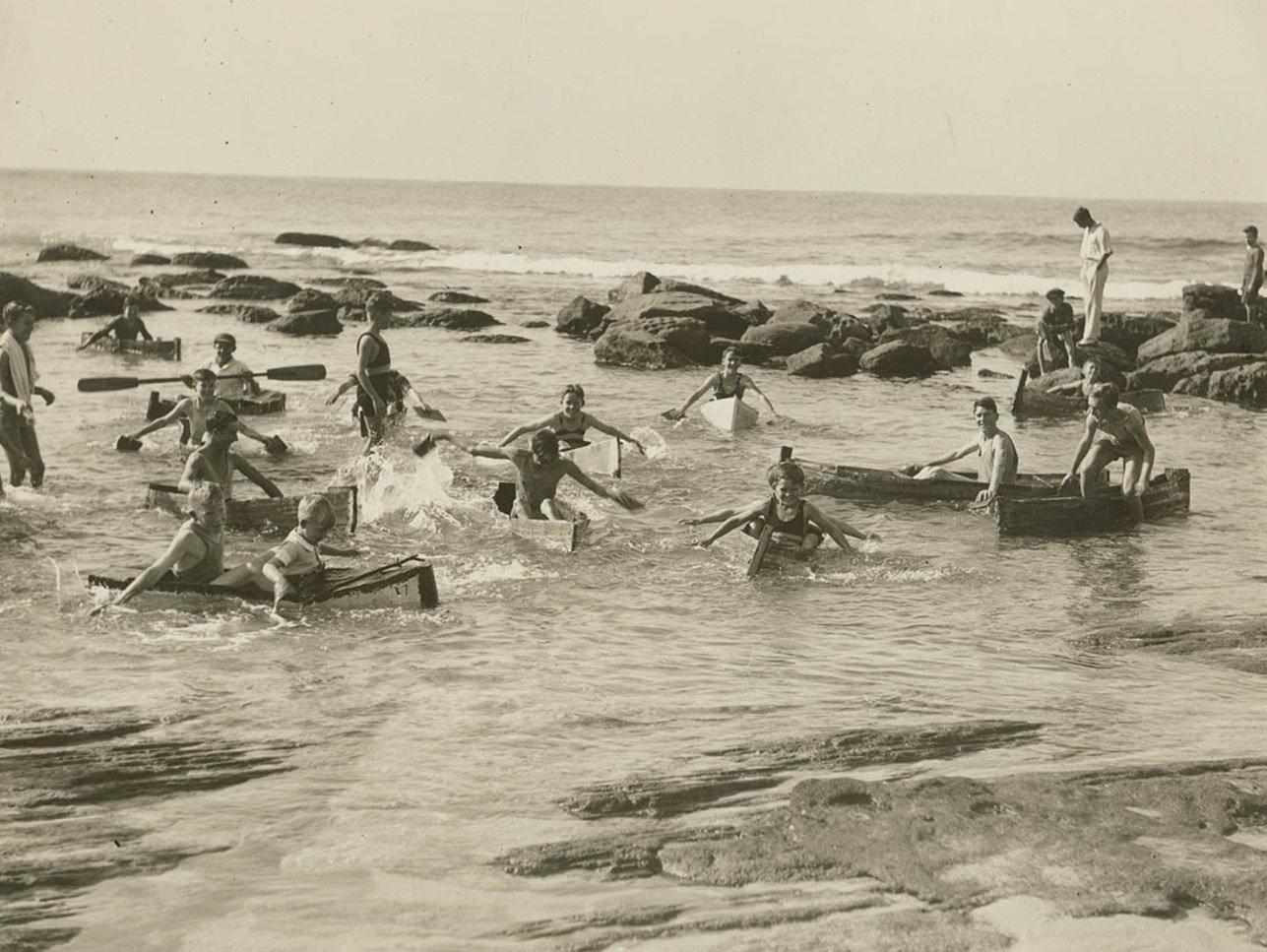 Men and boys swimming and playing in canoes in a beach rock pool.