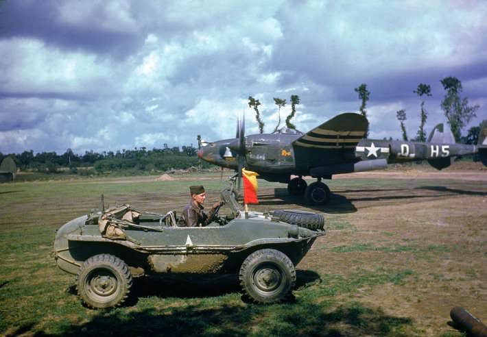 A P-38 fighter plane sits in the background as the pilot arrives in a captured German vehicle, France, 1944