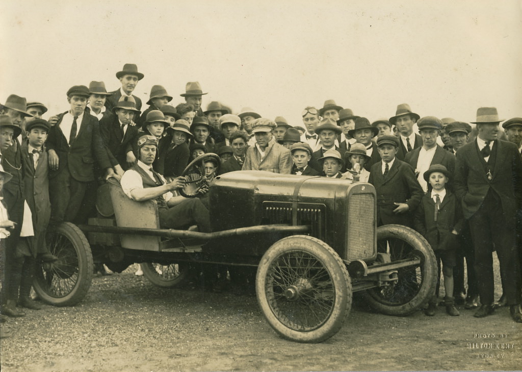 Man seated in a modified Overland racing car surrounded by crowd