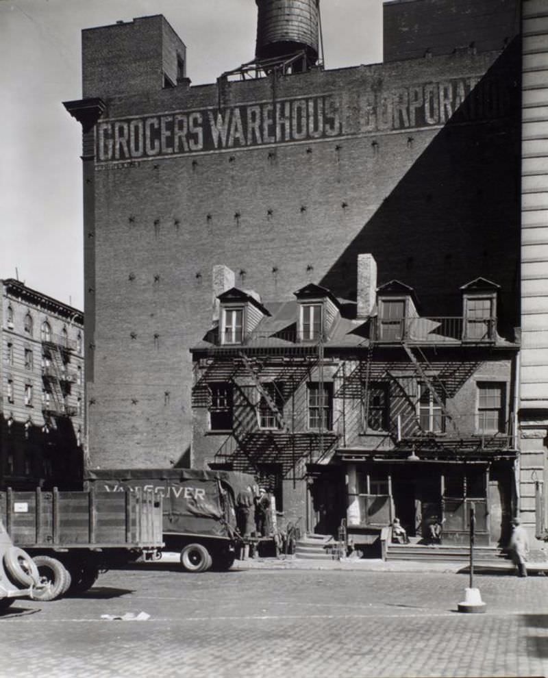 Broome Street no. 512-514, Manhattan. Two row houses are dwarfed by the Grocers Warehouse Corporation building behind it, trucks in the foreground.