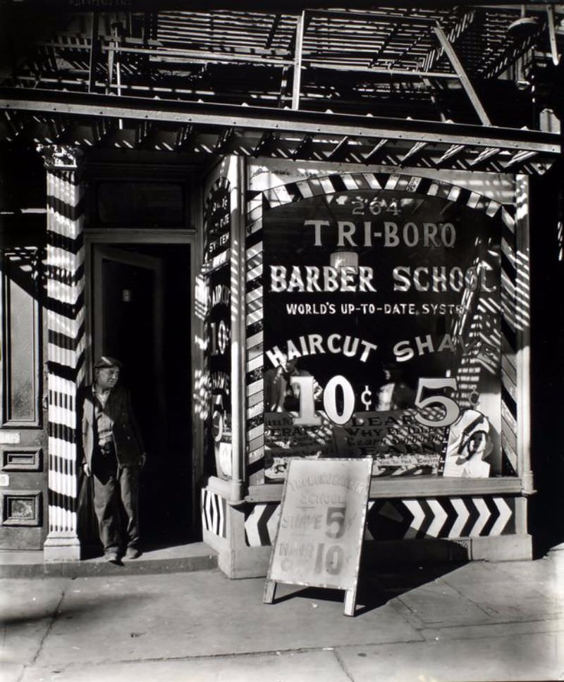 Tri-boro Barber School, 264 Bowery, Manhattan. Man leans on striped pilaster in doorway of barber school, men look out from behind advertisements painted on window, fire escape above.