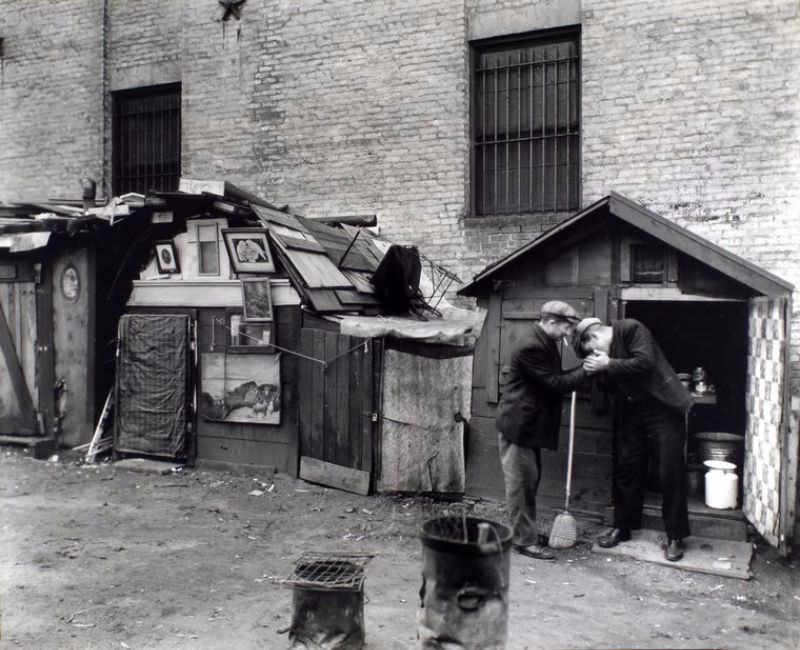 Men share a light in front of hut with open door, milk can and washtub inside, hut to left has pictures in frames adorning the outside of it.