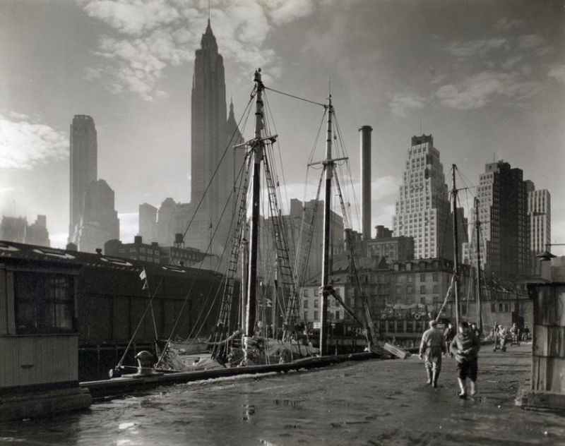 Fulton Street Dock, Manhattan skyline, Manhattan. Men walk on pier where sailing vessels are moored, skyline beyond.