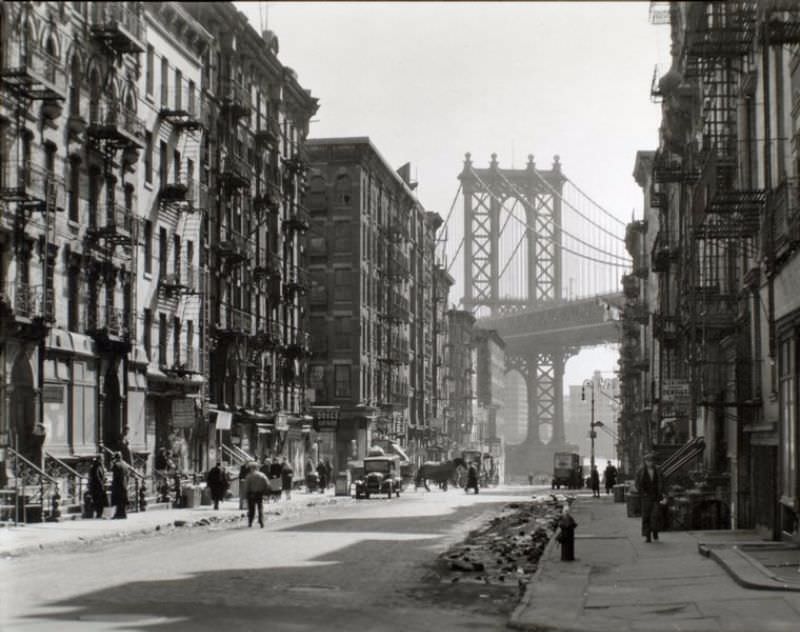 Looking down Pike Street toward the Manhattan Bridge, street half in shadow, rubble in gutters, some traffic.