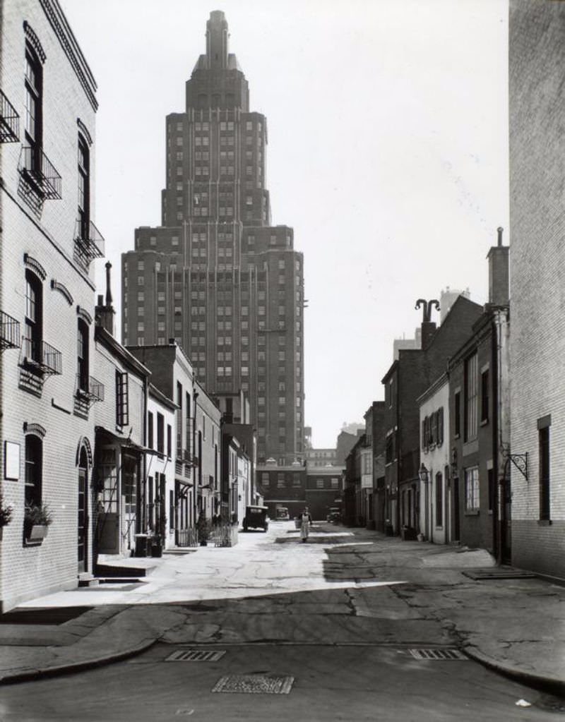 MacDougal Alley, between West 8th Street and Washington Square North, Manhattan. Man in apron with basket walks through alley with homes along it, 1 Fifth Avenue rises above.