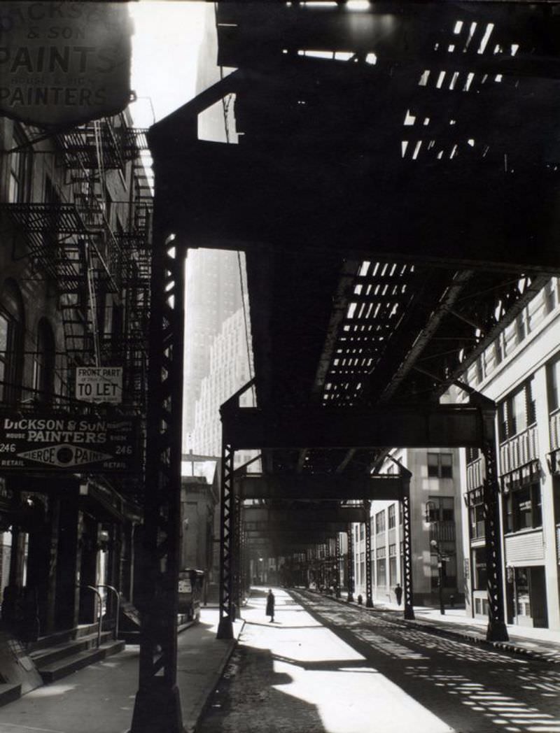 'El': 2nd & 3rd Avenue lines, looking W. from Second & Pearl St., Manhattan. Woman pauses below el tracks filling upper right, office buildings visible at left of tracks, painters shop in older building at left.