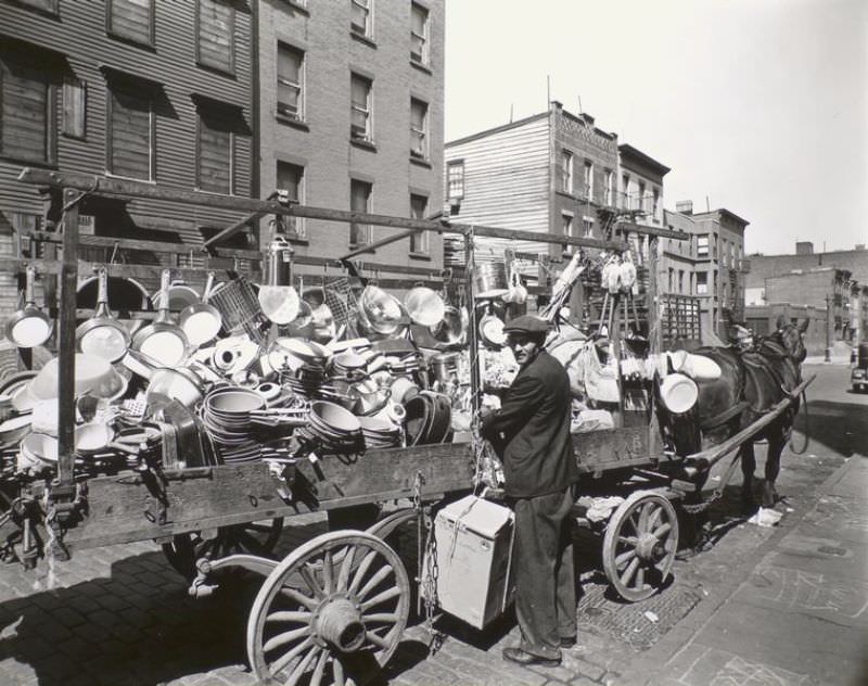 Tinker looks over his shoulder at camera while he ties box to wagon already loaded with pans, brushes, basins, etc.