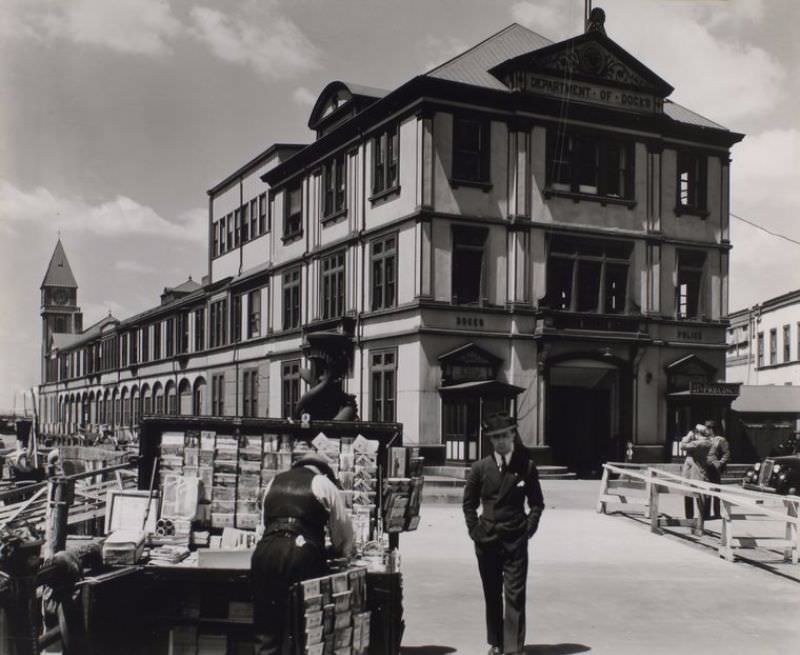 Long building wtih clock tower at far end housing Dept. of Docks and a Police station; man walks toward camera near newsstand in foreground.