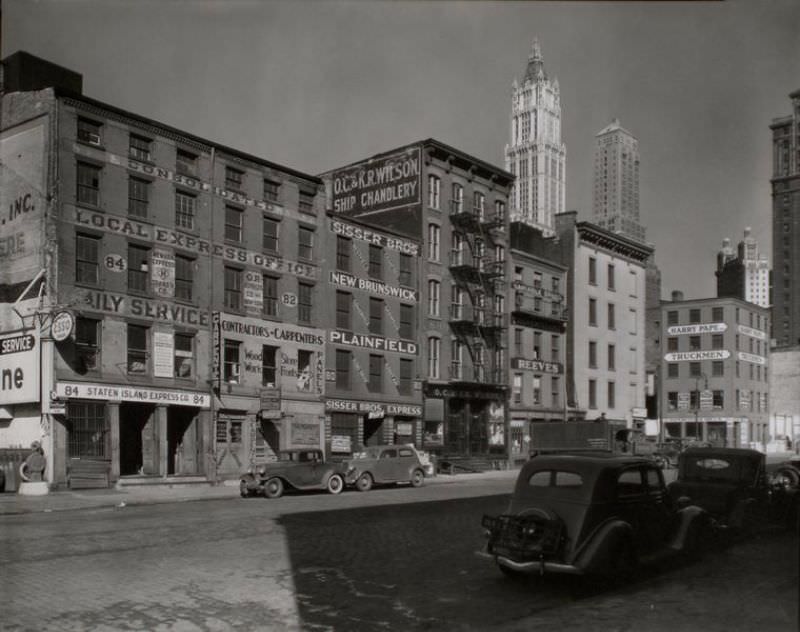 Dey Street between West and Washington Streets, Manhattan. Express companies, ships' outfitters and a contractor fill 4 and 5 story buildings along Dey St., tall towers beyond in upper right.