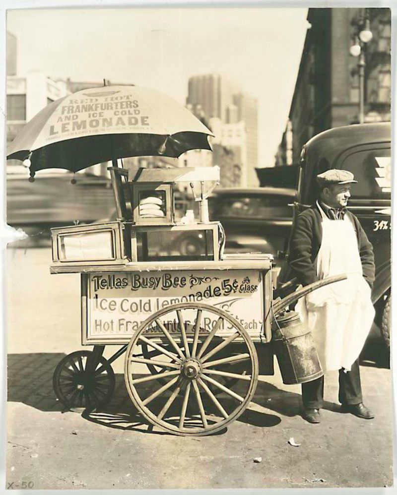 West St. and North Moore, Manhattan. Vendor stands next to his Tellas Busy Bee cart, advertising 'Red Hot Frankfurters and Ice Cold Lemonade' traffic a blur in the background.