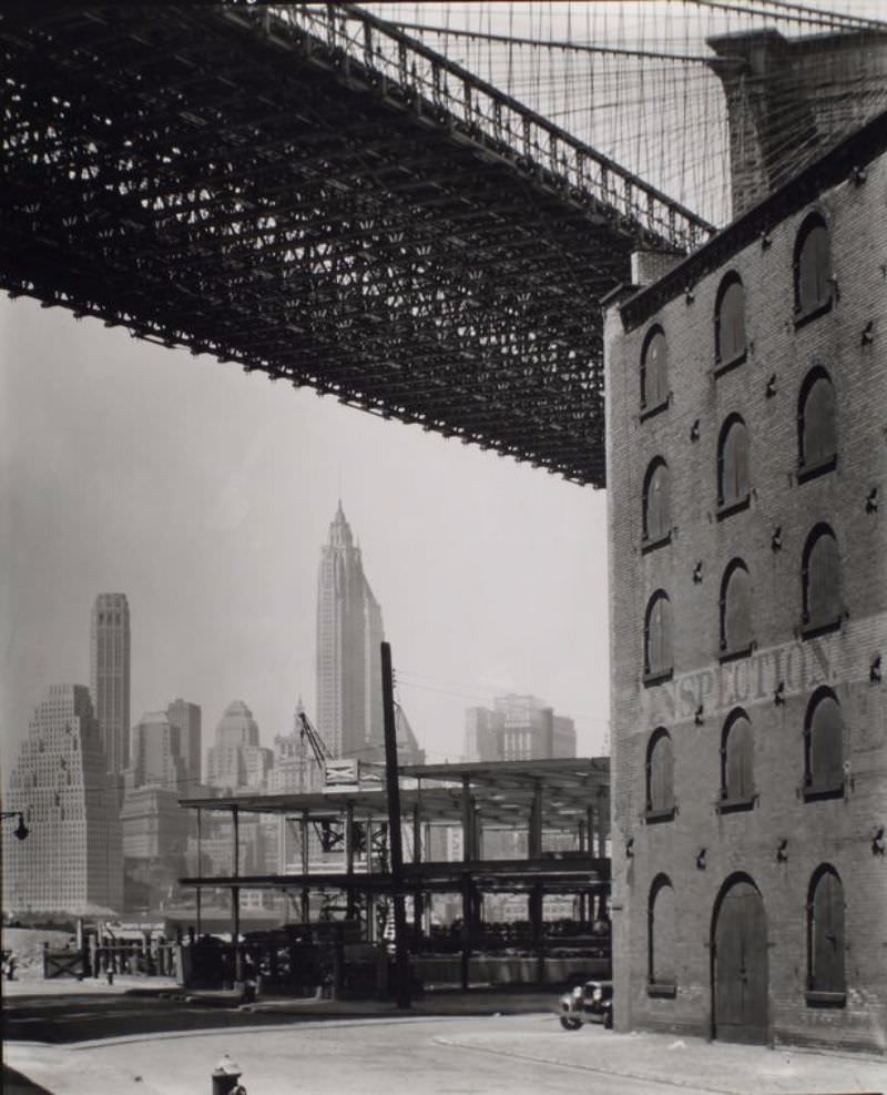 Steel frame building going up under the Brooklyn Bridge next to brick warehouse, Manhattan skyline in the distance.