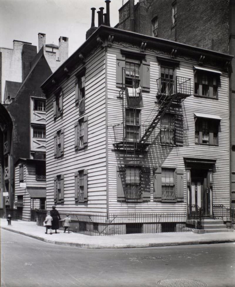 Frame House, Bedford and Grove Streets, Manhattan. Women and two children walk past corner house made with wood siding, shutters with crescent moons, top floor has rugs hanging out windows.