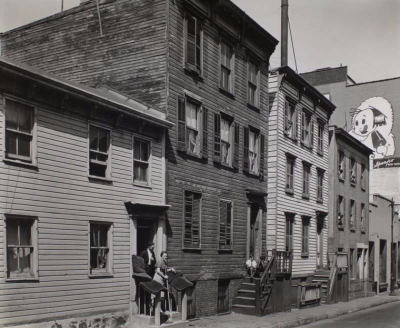 Talman Street, no. 57, Brooklyn. Man talks to woman with broom in hand on stoop of clapboard house, boys next door sit on stoop, Eskimo pie sign on building at right.