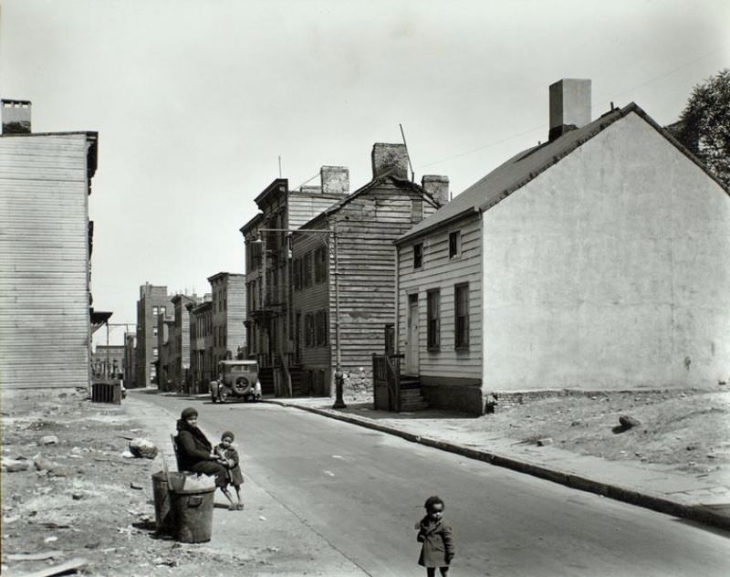 Talman Street, between Jay and Bridge street, Brooklyn. African American woman sits at street edge with two children, empty lots on either side of street, old 2 and 3 story clapboard houses further up.