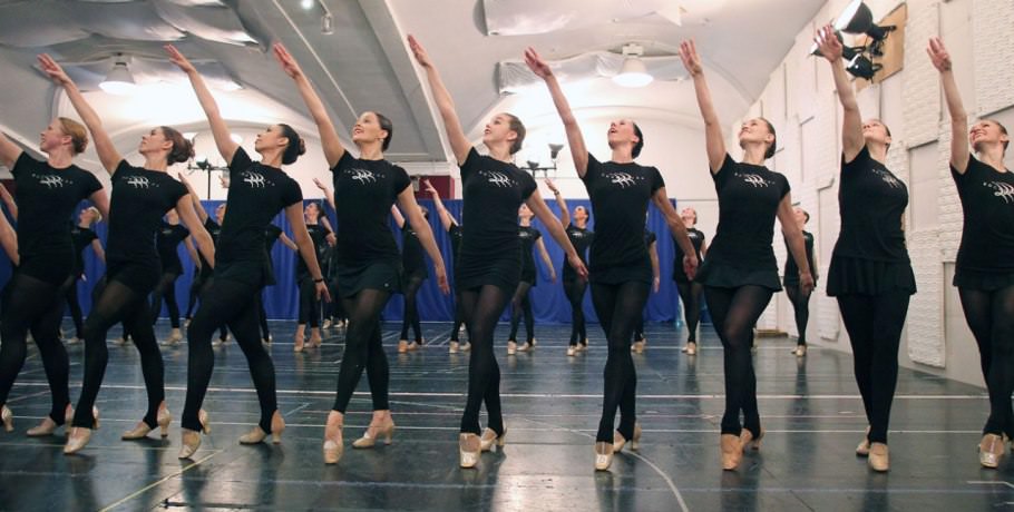 A handful of Rockettes strike a balletic pose at rehearsal, at which they all wear standard-issue black outfits and footless tights. The floor’s tape marks complex blocking for the Christmas Spectacular routines.