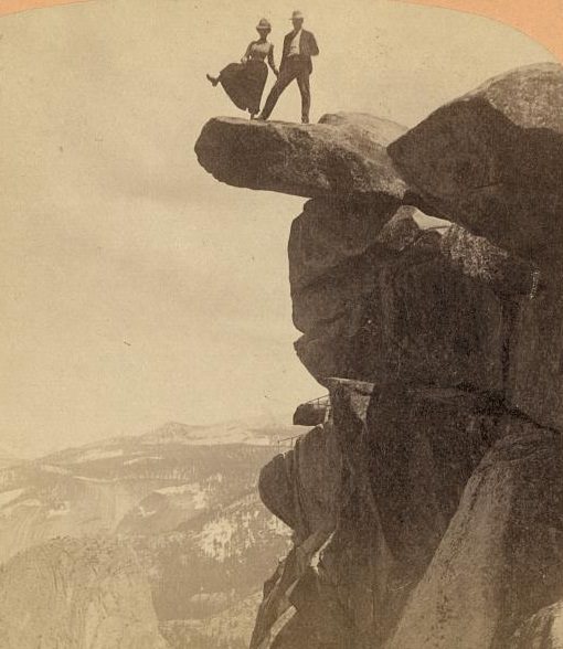 Man and woman standing on rock extending from top of cliff; woman is raising her right leg as though to step off.